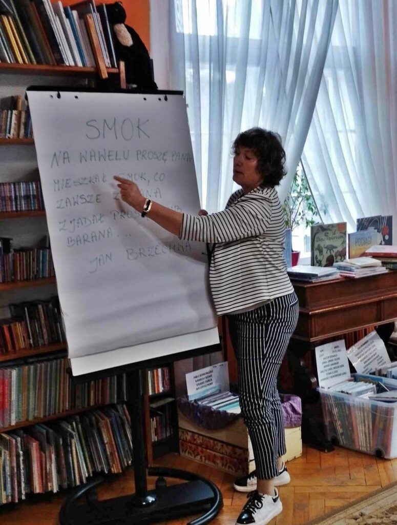 Bibliotekarka wskazuje palcem na przenośną tablicą. Na tablicy przyczepione białe kartki i tekst wierszyka Jana Brzechwy pt. Smok.