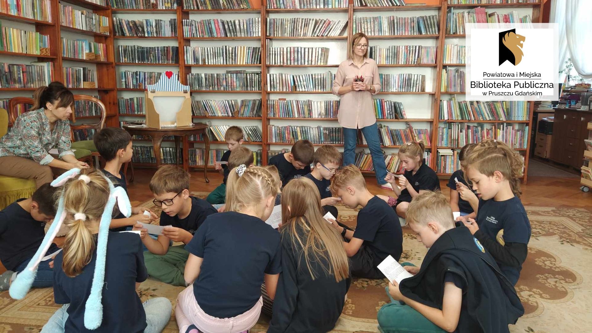 Dzieci siedzą na podłodze. Czytają teksty z kartek. Po lewej siedzi nauczycielka. Za uczniami stoi bibliotekarka i trzyma w ręce pisaki. W tle regały z książkami.
