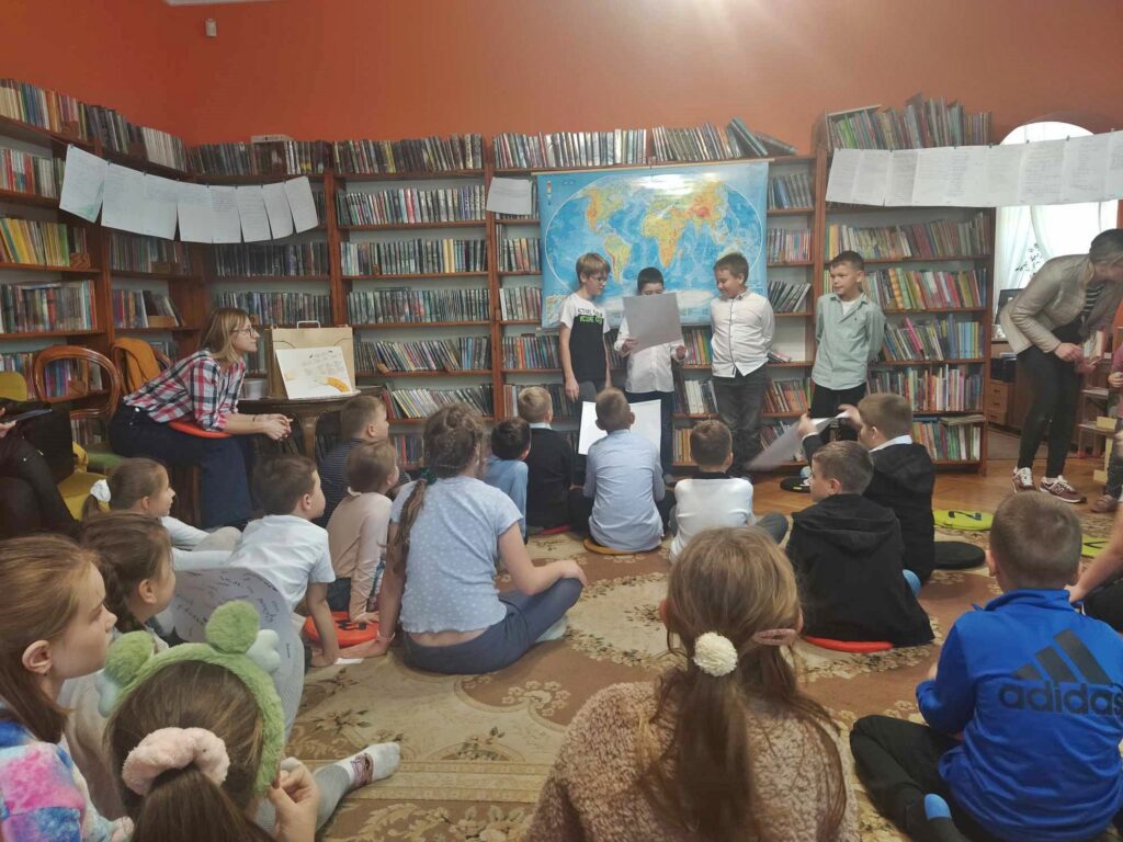 Czterech chłopców stoi przed grupą dzieci siedzących na podłodze. Jeden z chłopców trzyma kartkę. Za nimi wisi mapa świata. Po lewej siedzi bibliotekarka. W tle regały z książkami.