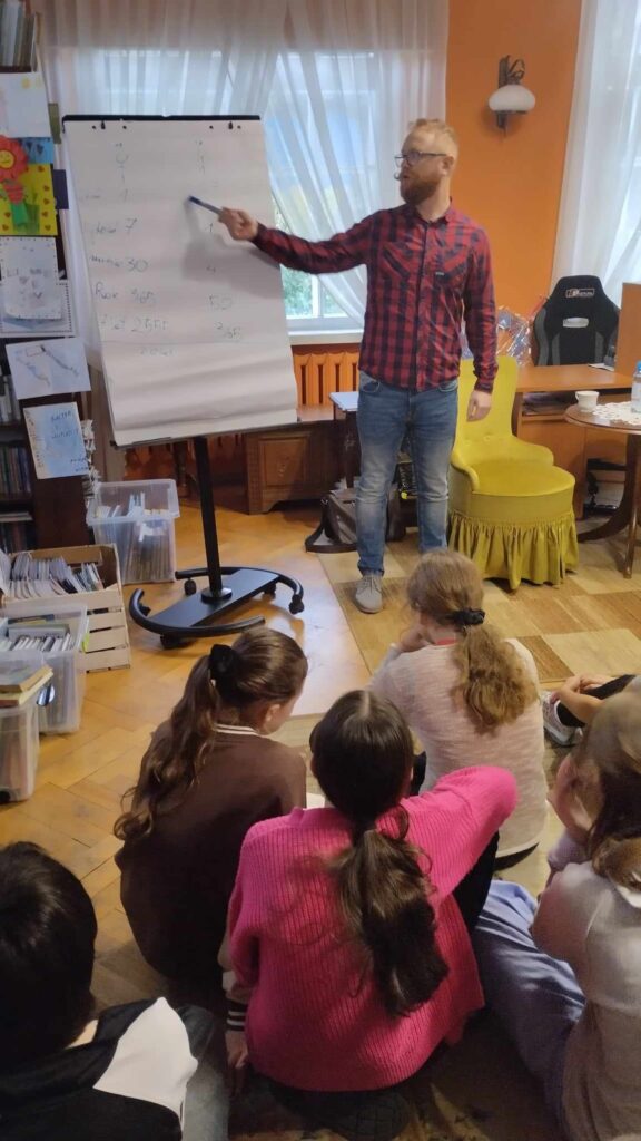 Na dywanie siedzi młodzież. Młodzież patrz na stojącego przed nimi Krzysztofa Piersę, który wskazuje flamastrem na zapisaną tablicę przenośną.