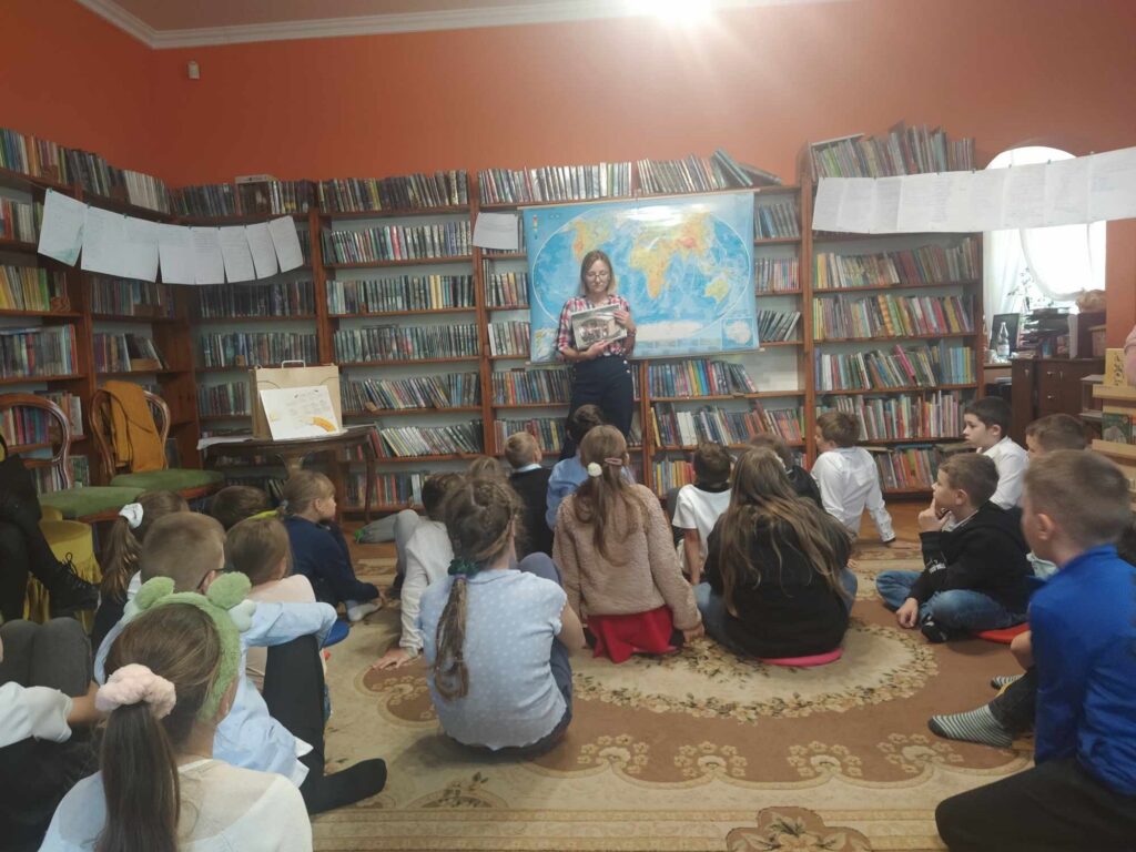 Dzieci siedzą na podłodze i patrzą w kierunku bibliotekarki, która pokazuje ilustrację. Za bibliotekarką wisi mapa świata. W tle regały z książkami.