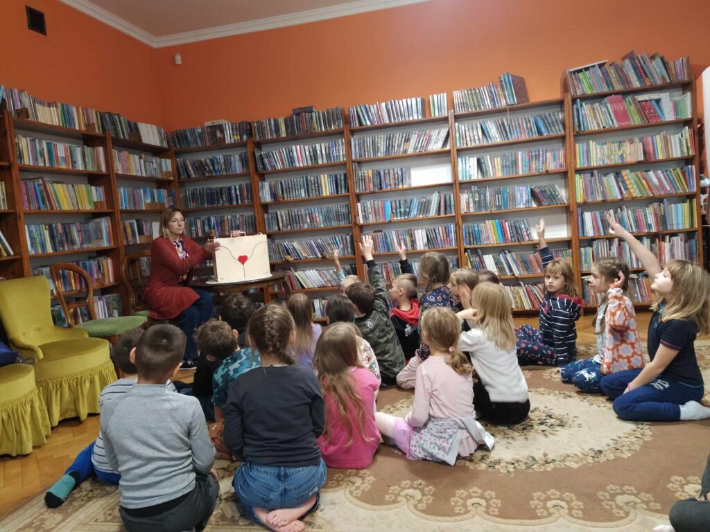 Dzieci siedzą na podłodze, niektóre z nich zgłaszają się. Bibliotekarka siedzi przy stoliku, na którym stoi teatrzyk kamishibai. W tle regały z książkami.