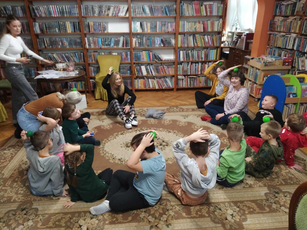 Dzieci wraz z paniami siedzą na podłodze i masują głowę piłkami sensorycznymi. W tle regały z książkami.