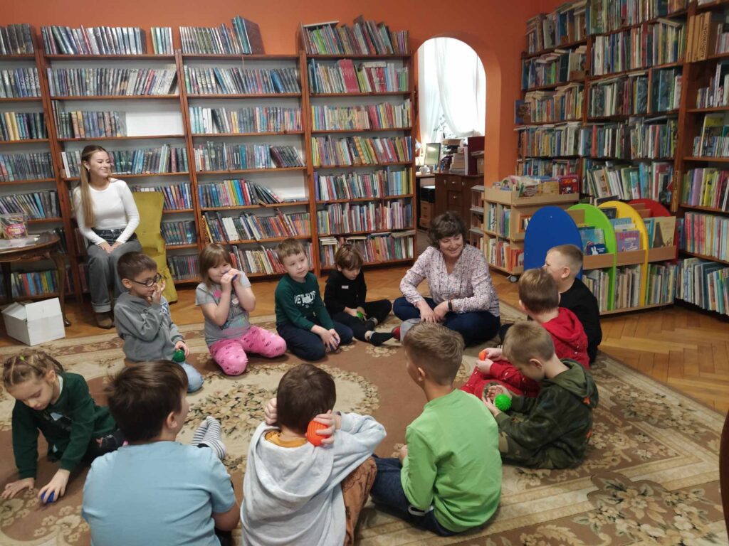Dzieci wraz z bibliotekarką siedzą na podłodze. W rękach mają piłki sensoryczne. W tle regały z książkami.