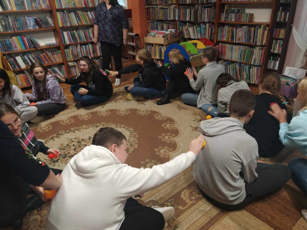 Dzieci siedzą w okręgu i wykonują sobie wzajemnie masaż pleców piłkami sensorycznymi. W tle regały z książkami.