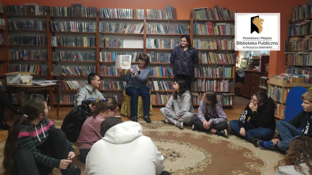 Dzieci siedzą na podłodze. Bibliotekarka siedzi na krześle i pokazuje ilustrację w książce. Po prawej stoi druga bibliotekarka. W tle regały z książkami.