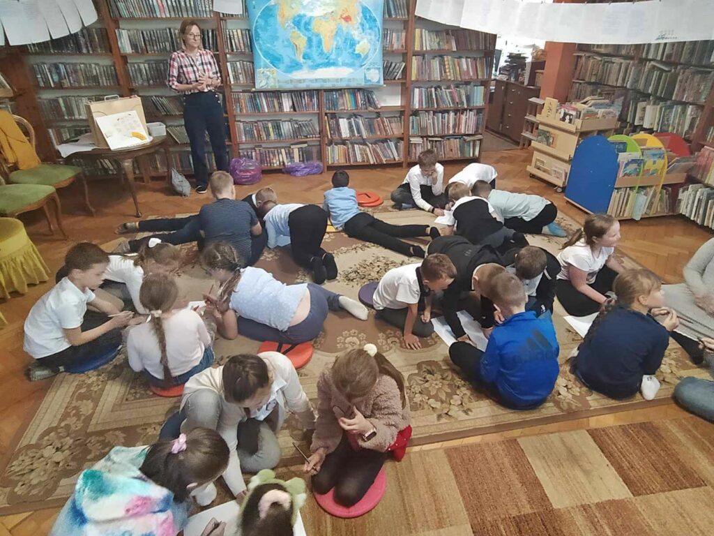 Dzieci siedzą i leżą na podłodze. Piszą na kartkach. Bibliotekarka stoi przed nimi. W tle wisi mapa świata i znajdują się regały z książkami.