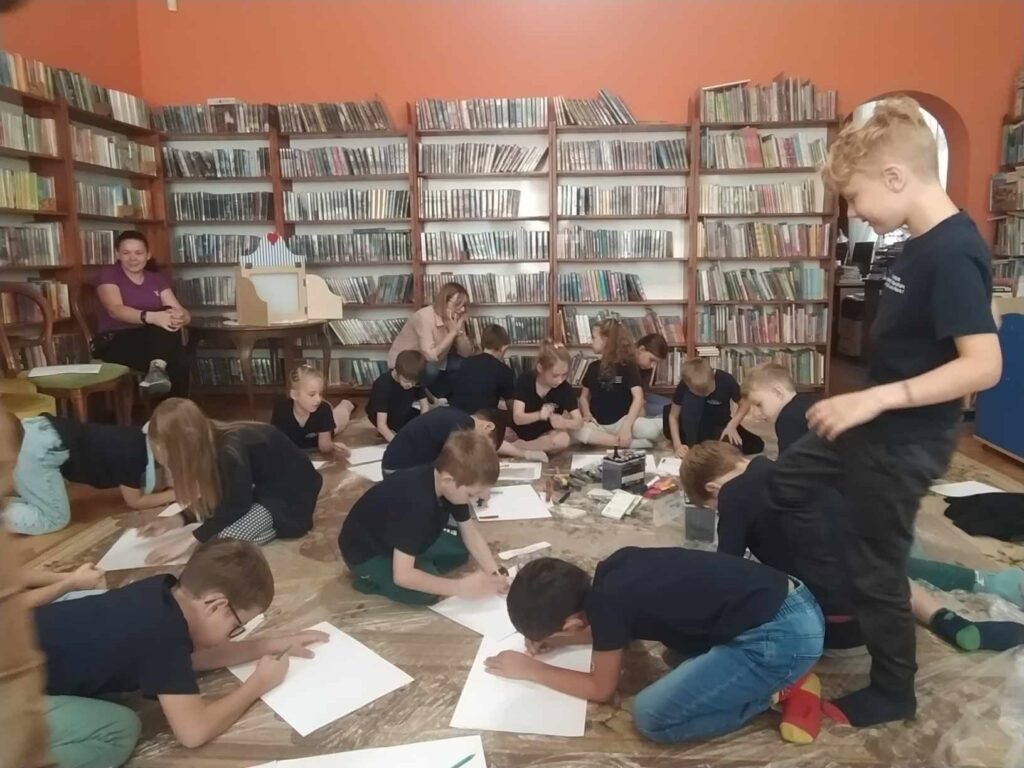 Dzieci siedzą na podłodze i rysują. Jeden z chłopców stoi i przygląda się. Po lewej siedzi nauczycielka. Za dziećmi siedzi bibliotekarka. W tle regały z książkami.