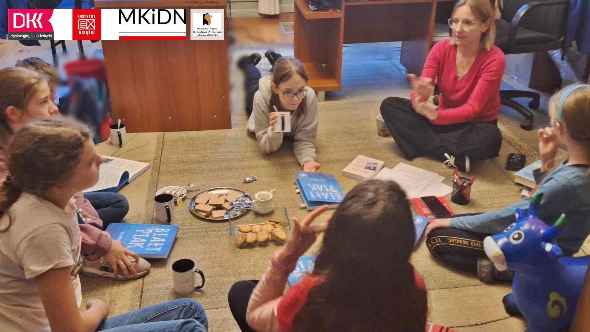 Kilkoro dzieci siedzi na dywanie w okręgu. Między nimi siedzi moderatorka klubu, Joanna Łabasiewicz. Pośrodku leżą ciastka, książki oraz stoją kubki z napojem.