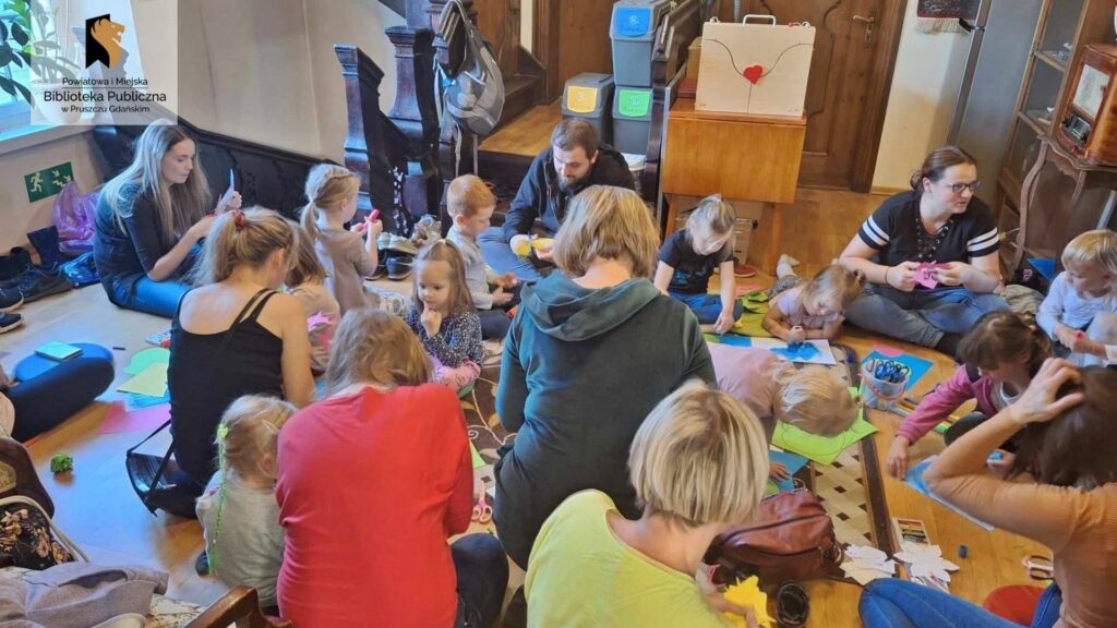 Dizeic oraz rodzice uczestniczący w spotkaniu Klubu książki dla przedszkolaka. Wszyscy siedzą na dywanie, dorośli wycinają, a dzieci malują mazakami oraz naklejają nenufary na kartki.