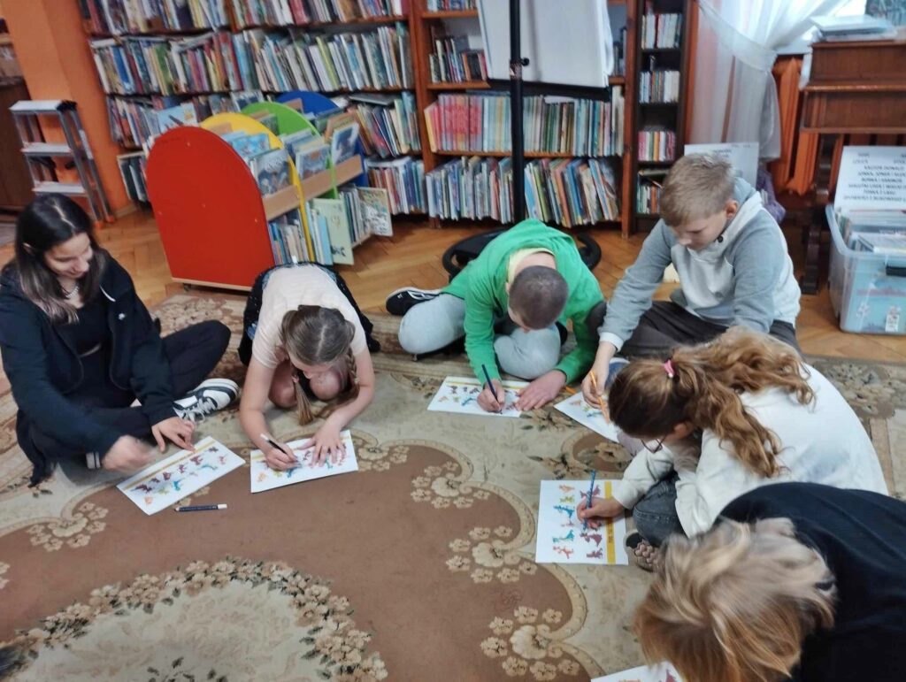 Na dywanie w sali bibliotecznej siedzą dzieci. Przed dziećmi leżą kartki, na których widnieją sylwetki smoka. Niektóre dzieci trzymają w rekach mazaki.
