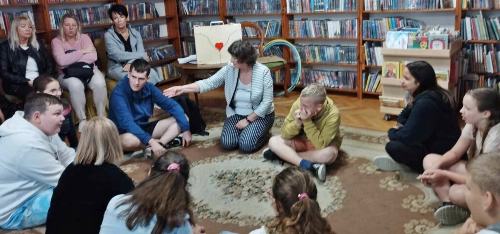 Na dywanie w sali bibliotecznej siedzą dzieci. Między nimi siedzi bibliotekarka, Anna Krawycińska. Za nimi na stoliku stoi drewniana skrzynka-teatrzyk kamishibai.
