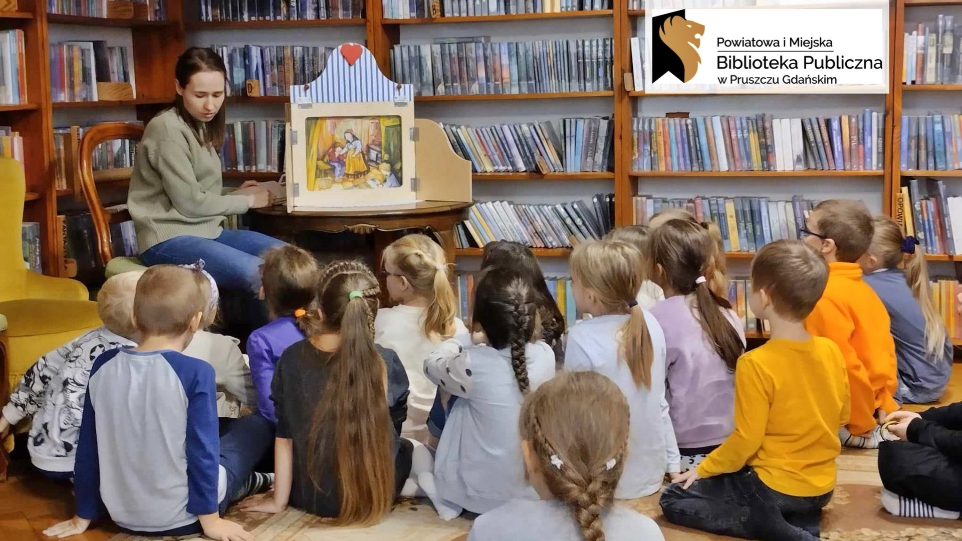 Na dywanie w bibliotece siedzi duża grupa dzieci. Dzieci patrzą na bibliotekarkę, która siedzi obok otwartego teatrzyku. W teatrzyku ilustracja z bajki Jaś i Małgosia: 2 dzieci i 1 osoba dorosła.