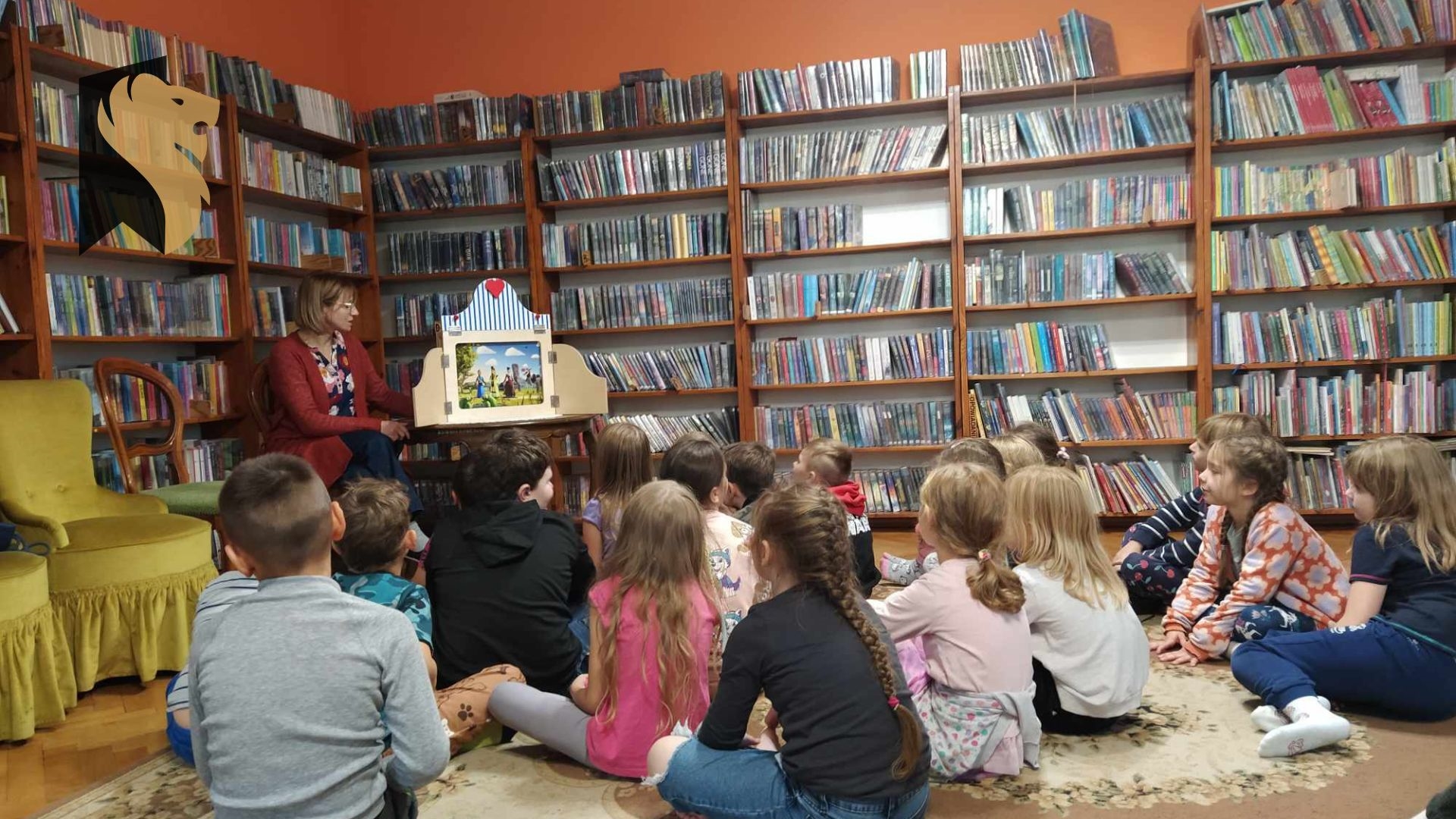 Dzieci siedzą na podłodze i patrzą w stronę teatrzyku kamishibai, który stoi na stoliku. Po lewej siedzi bibliotekarka i czyta. W tle regały z książkami.