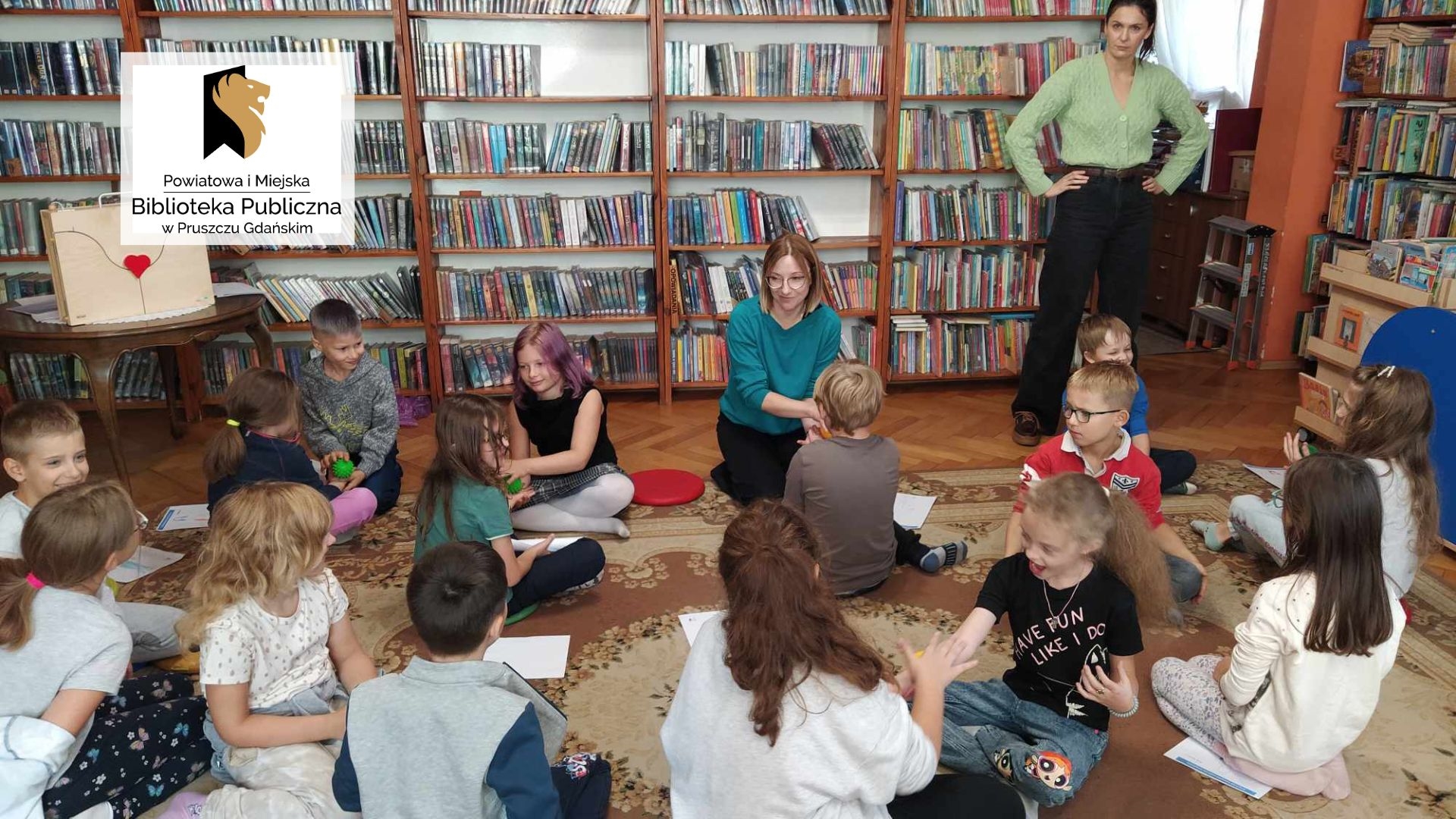 Dzieci wraz z bibliotekarką siedzą na podłodze. Masują sobie wzajemnie ręce piłkami sensorycznymi. Nauczycielka stoi za nimi. W tle regały z książkami.
