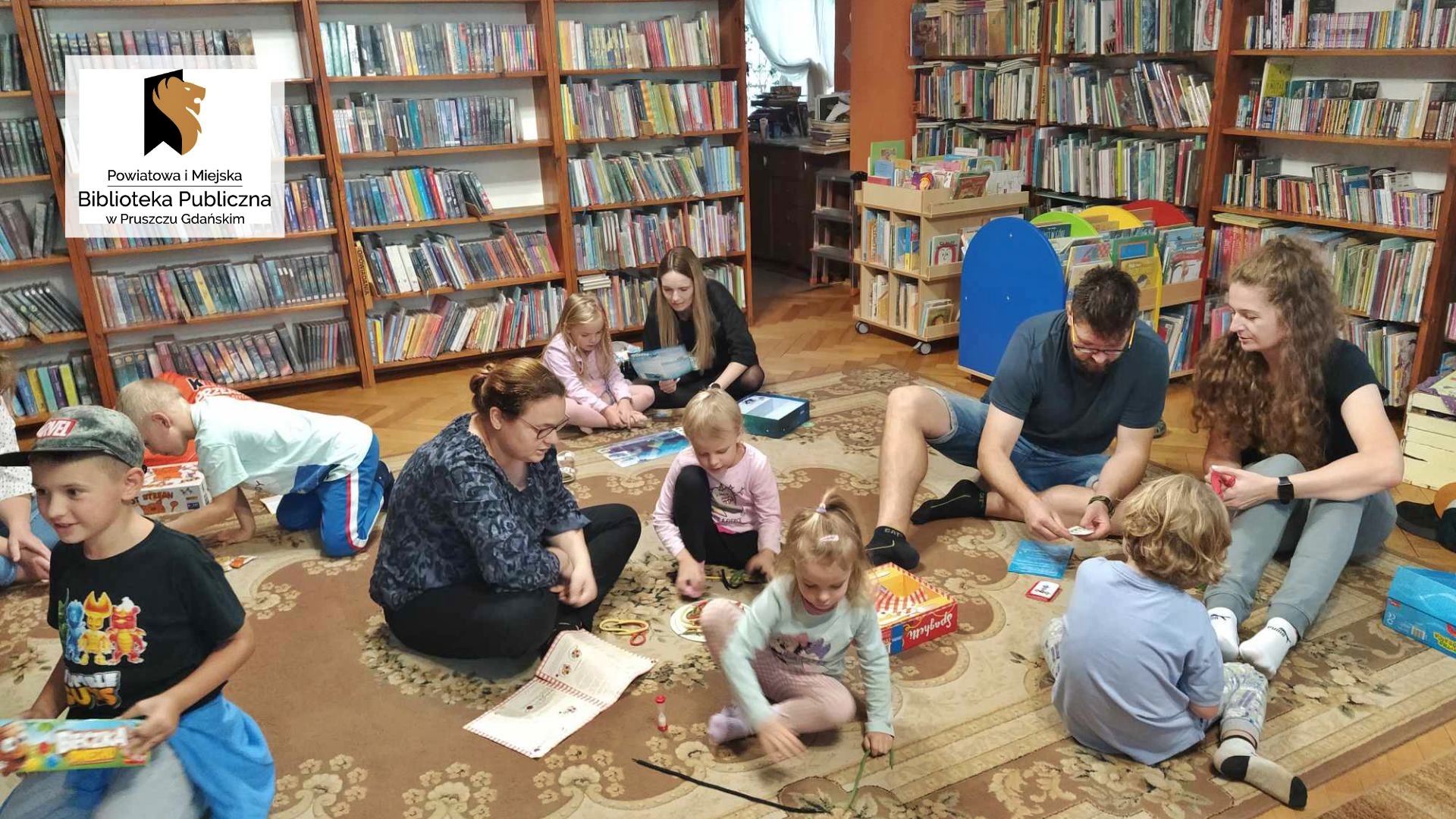 Dzieci i dorośli siedzą na podłodze. Przed nimi leżą różne gry. W tle regały z książkami.