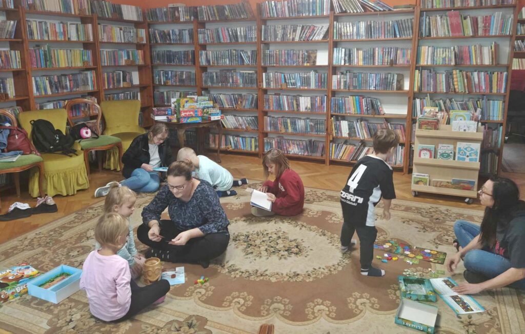 2 Dzieci i dorośli siedzą na podłodze. Jeden chłopiec stoi. Przed nimi znajdują się gry. Jedna z dziewczynek czyta książkę. W tle regały z książkami.