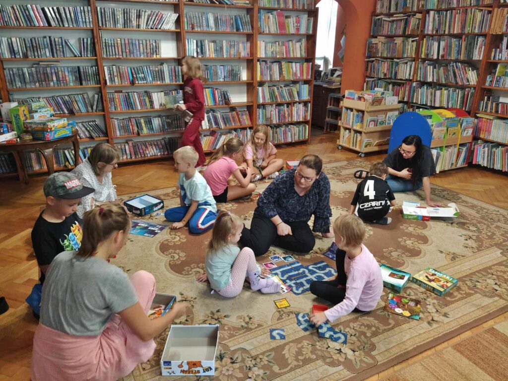 Dzieci wraz z dorosłymi siedzą na podłodze, przed nimi leżą różne gry. Jedna dziewczynka przechodzi przez pomieszczenie. Po lewej znajduje się stolik, na którym leżą gry planszowe. W tle regały z książkami.