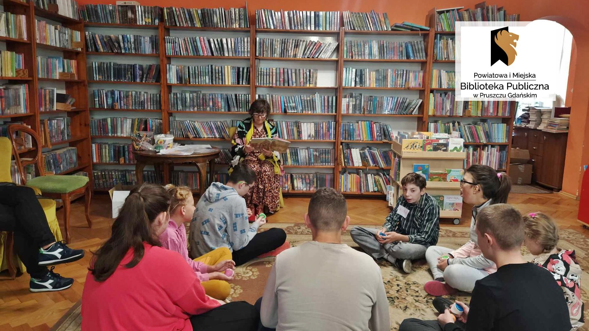 Dzieci siedzą w okręgu na podłodze. Pani bibliotekarka siedzi i czyta książkę. W tle stolik z materiałami plastycznymi i regały z książkami.