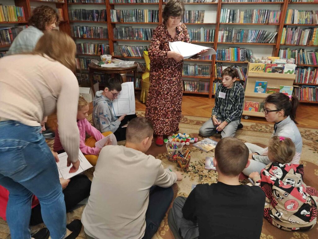 Dzieci siedzą na podłodze. Panie rozdają im kartki. Przed nimi leżą kredki i pisaki. Na podłodze leżą kolorowe piłki. W tle regały z książkami.