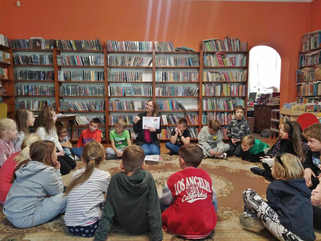 Dzieci wraz z panią bibliotekarką siedzą w okręgu na podłodze. Pani trzyma planszę z grafiką i tekstem. W tle regały z książkami. 