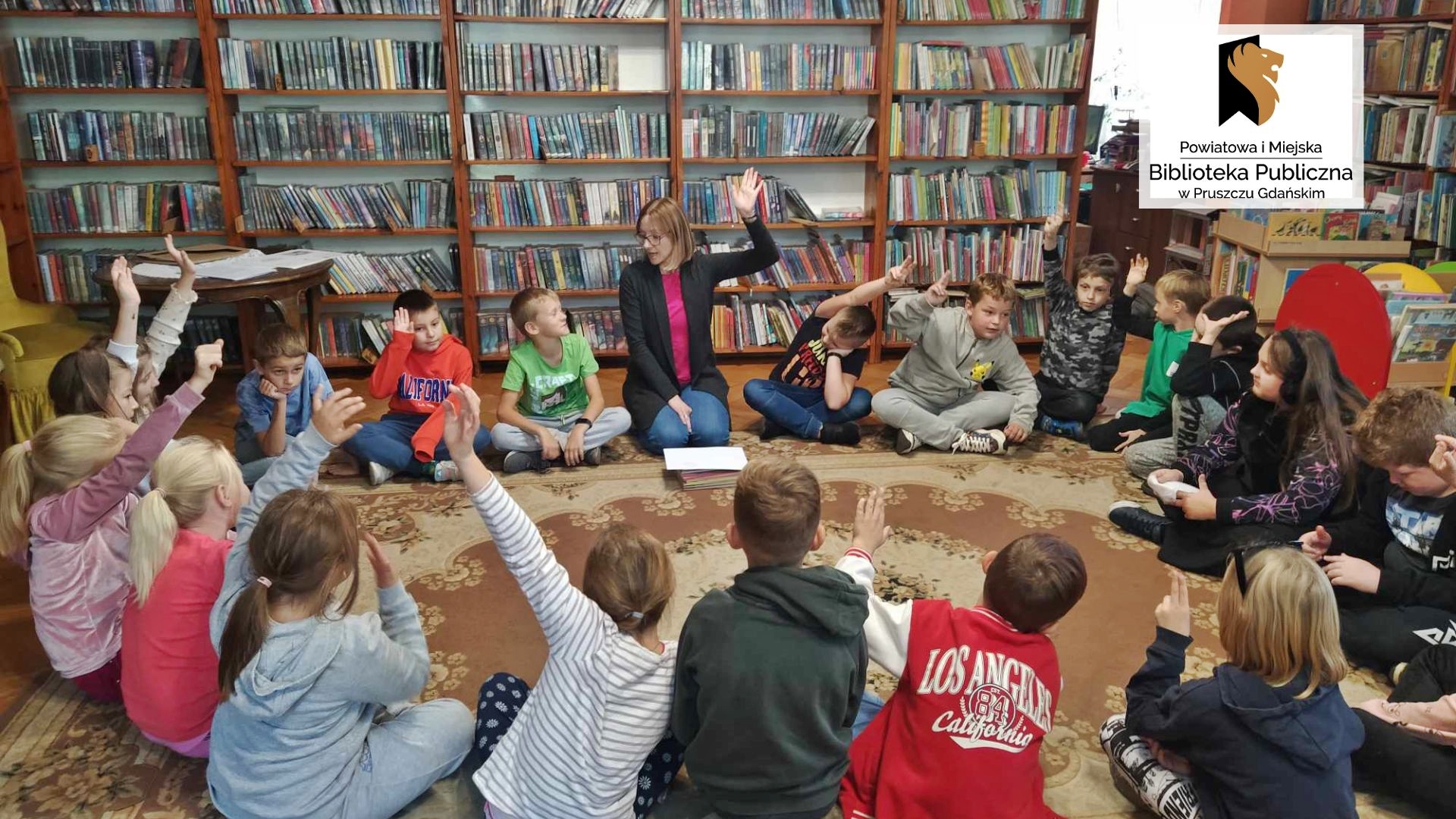 Dzieci wraz z panią bibliotekarką siedzą na podłodze w okręgu. Pani i niektóre z dzieci zgłaszają się gestem dłoni. W tle regały z książkami.