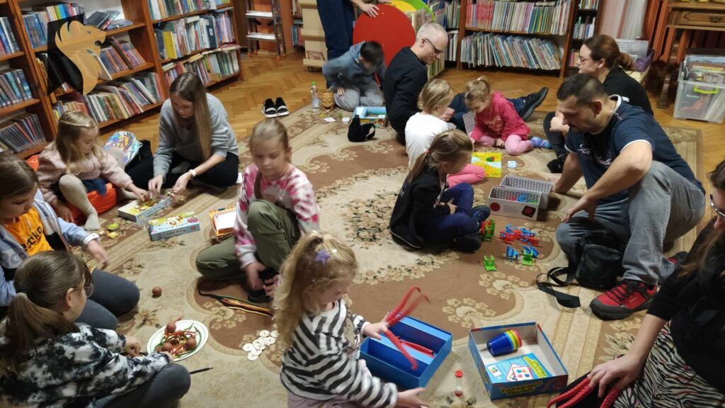Dzieci i dorośli siedzą na podłodze i grają w gry planszowe. W tle regały z książkami.