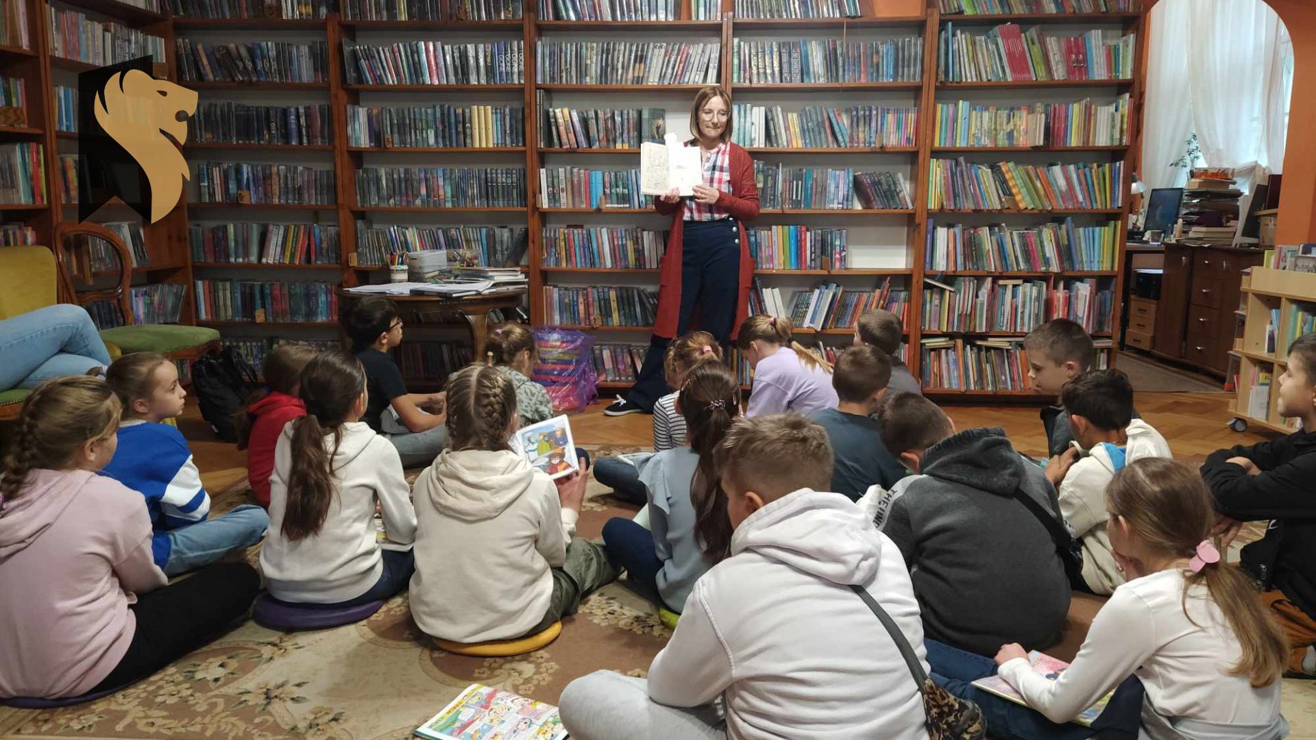 Dzieci siedzą na podłodze i patrzą w kierunku bibliotekarki. Bibliotekarka trzyma przed sobą otwartą książkę i pokazuje ją dzieciom. W tle stolik z materiałami plastycznymi i regały z książkami.