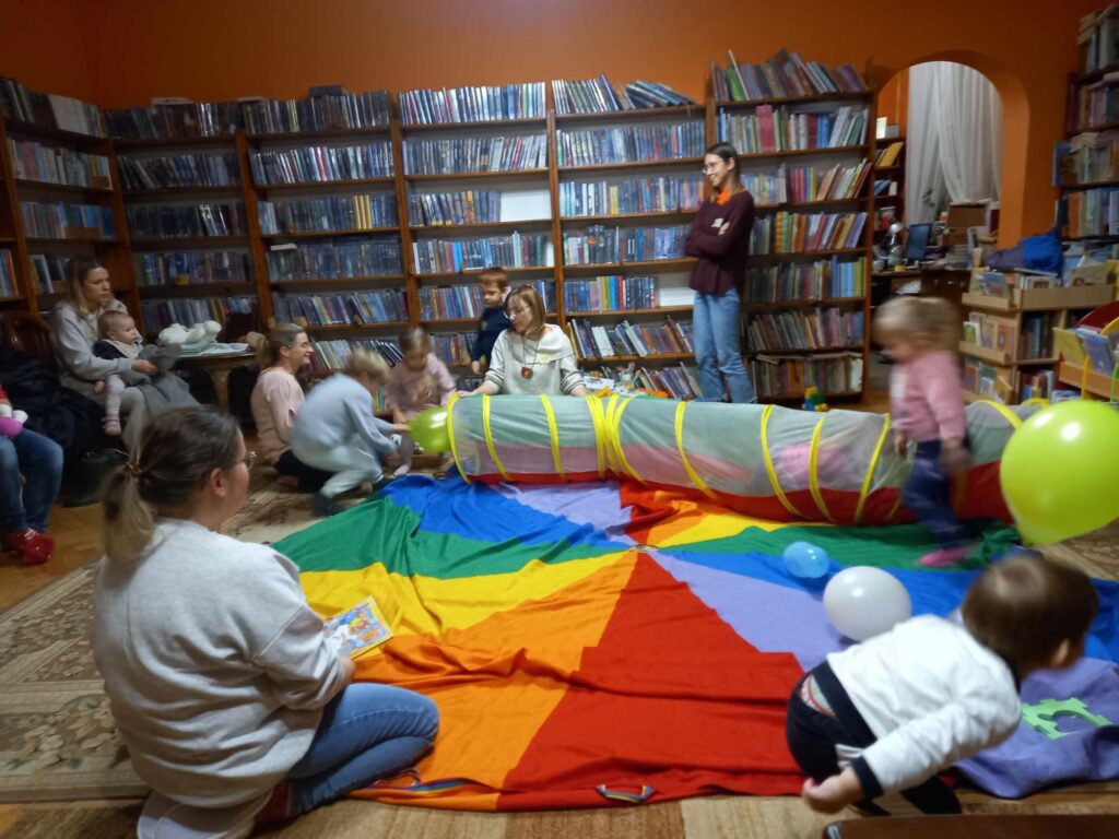Na dywanie w bibliotece rozłożona kolorowa chusta animacyjna. Na chuście leży kolorowy tunel, do którego wychodzą jedną stroną, a w drugą wychodzą dzieci. Tunel trzyma bibliotekarka. Obok chusty stoją i siedzą rodzice.