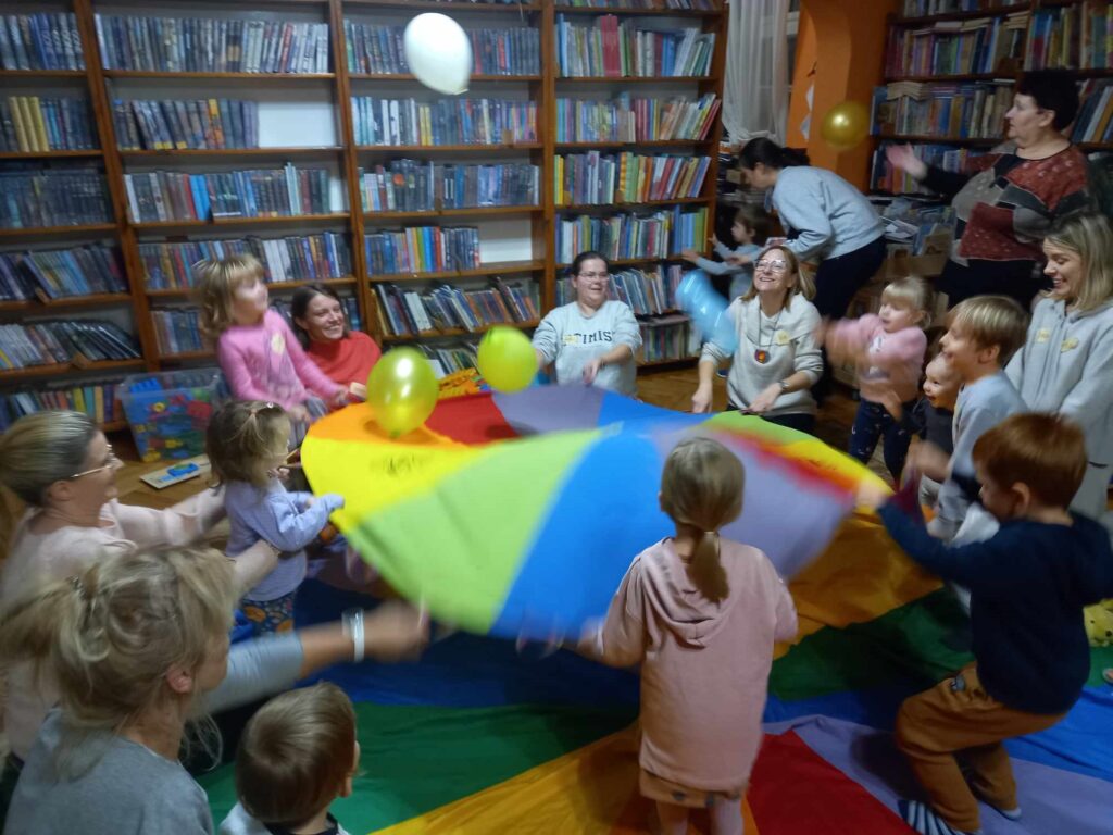 Na kolorowej chuście animacyjnej stoją małe dzieci. Dzieci wraz z bibliotekarką i rodzicami potrząsają mniejszą chustą animacyjną, na której leżą kolorowe balony. Wszyscy się śmieją.
