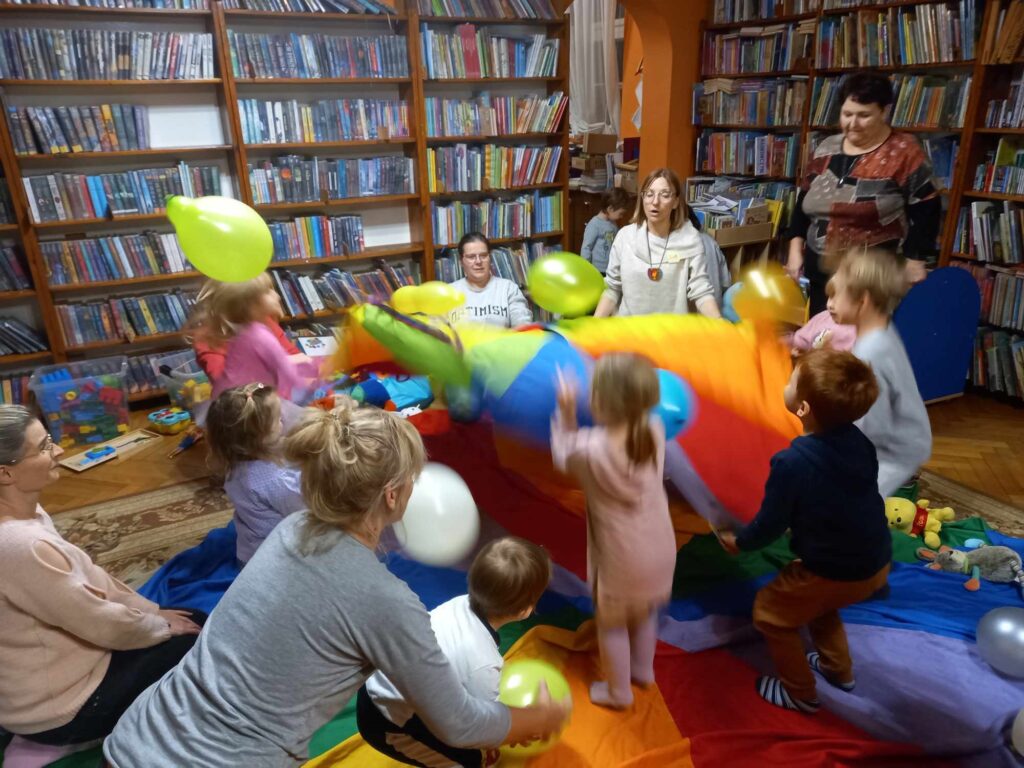 Na kolorowej chuście animacyjnej stoją małe dzieci. Dzieci wraz z bibliotekarką i rodzicami potrząsają mniejszą chustą animacyjną, na której leżą kolorowe balony.