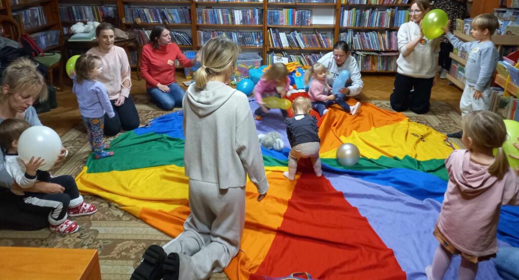 Na dywanie w wypożyczalni dla dzieci leży chusta animacyjna. Na chuście stoi 3 małych dzieci oraz leżą balony. Wkoło chusty siedzą na dywanie rodzice i dzieci.