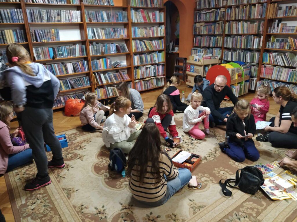 Dzieci i dorośli siedzą na podłodze i grają w gry planszowe. W tle regały z książkami.