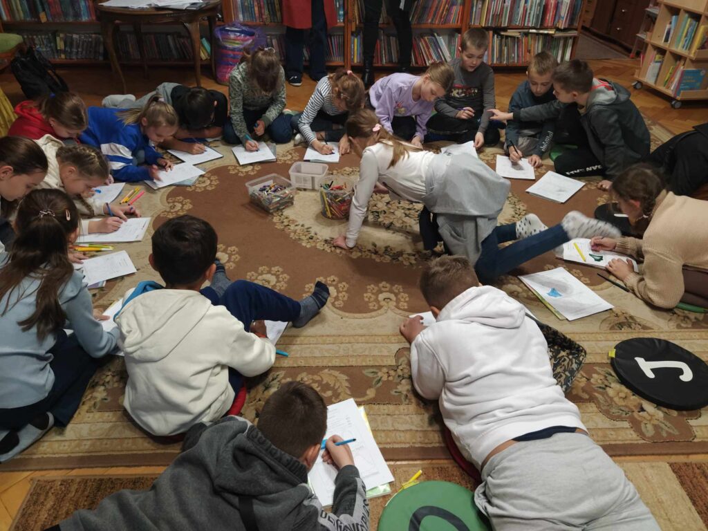 Dzieci siedzą na podłodze. Rysują na kartkach, które mają przed sobą. Jedna dziewczynka sięga do środka okręgu po kredki. W tle regały z książkami.