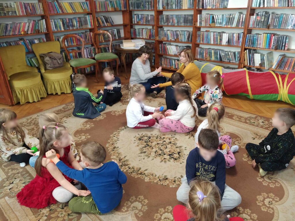 Dzieci wraz z paniami siedzą na podłodze i masują sobie wzajemnie ręce przy pomocy piłek sensorycznych. W tle regały z książkami.