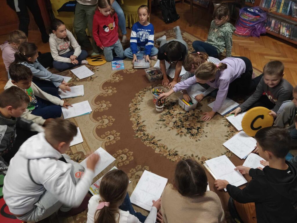 Dzieci siedzą na podłodze. Przed sobą mają kartki. Kilkoro z dzieci sięga po kredki.