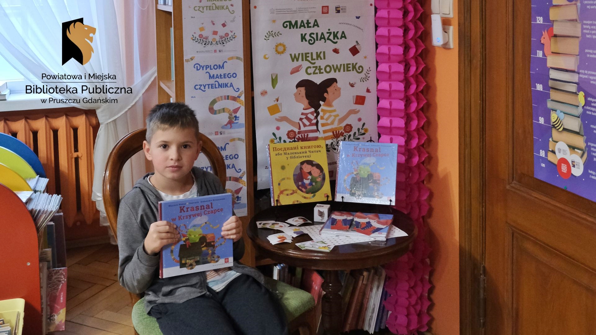 Chłopiec siedzi na krześle i trzyma w rękach książkę pt. Krasnal w Krzywej Czapce. Obok na stoliku wyeksponowane te same książki, jedna z nich w języku ukraińskim. Obok książek: papierowa kostka, naklejki i plakietka do zbierania nalepek. Za stolikiem plakat z napisem: mała książka-wielki człowiek.