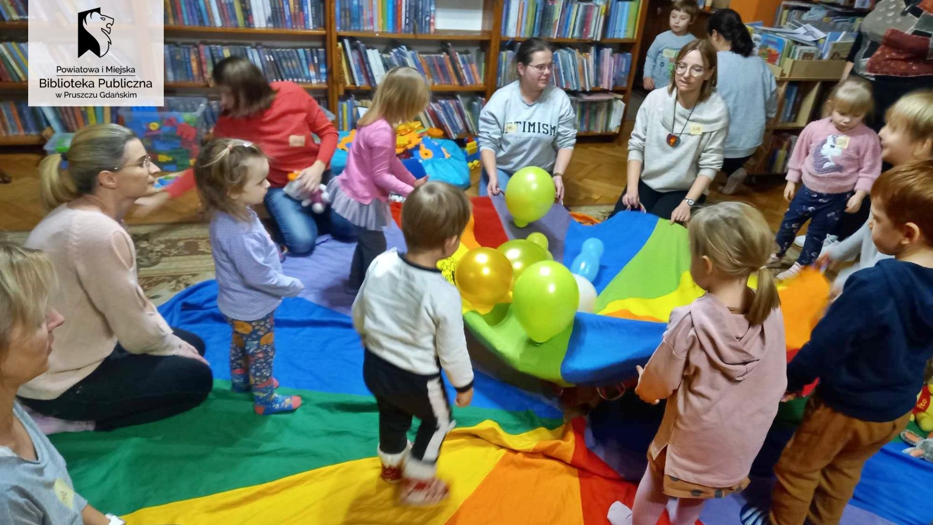 Na dywanie rozłożona kolorowa chusta Klanzy. Na środku chusty leżą kolorowe balony: żółte, zielone, białe, niebieskie, niebieskie. Obok balonów stoją małe dzieci. Kilkoro dorosłych trzyma chustę za rogi, a niektórzy na niej siedzą.