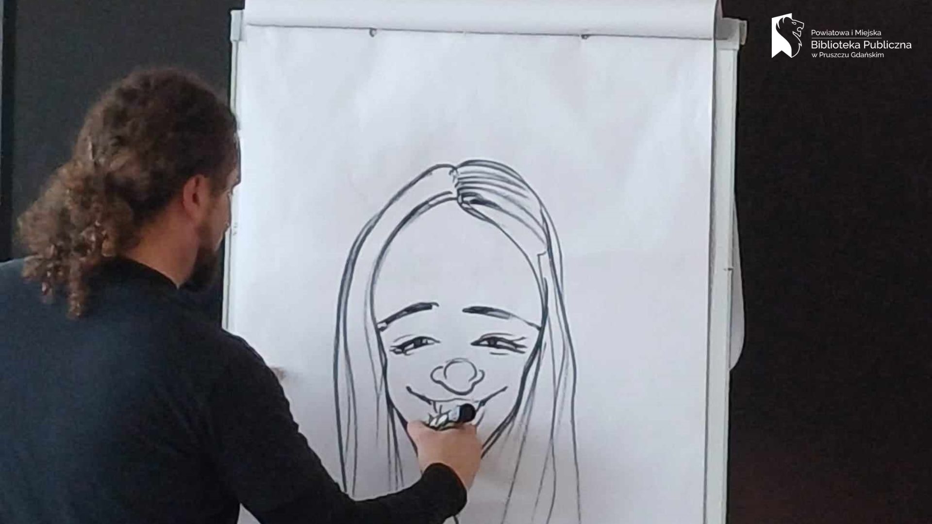 Szymon Teluk prezentuje narysowaną przez siebie, na białym papierze, twarz kobiety. Kobieta ma długie włosy i uśmiecha się.