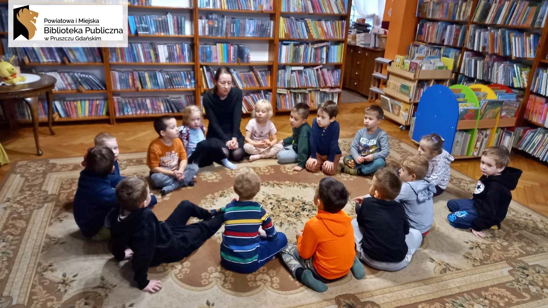 Na dywanie w wypożyczalni dla dzieci siedzą przedszkolaki. Między dziećmi siedzi bibliotekarka i coś mówi.