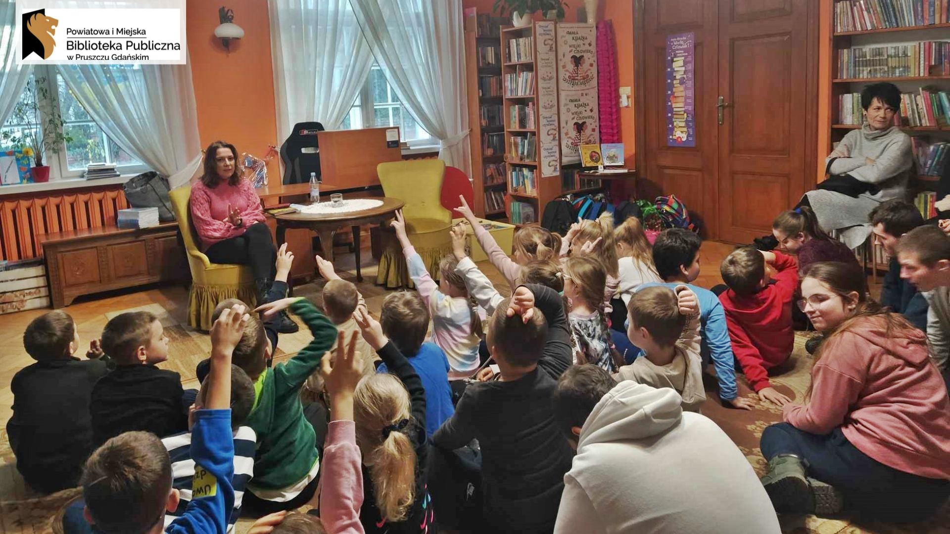 Dzieci siedzą na podłodze i zgłaszają się gestem dłoni. Przed nimi siedzi na fotelu Magda Bielicka.
