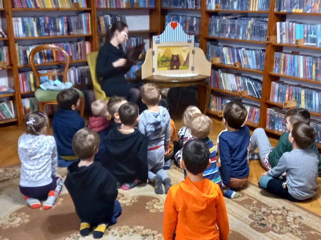 Dzieci siedzą na podłodze i patrzą w kierunku bibliotekarki, która mówi i wykonuje gest dłonią. Po lewej, na stoliku, stoi teatrzyk kamishibai, w środku znajduje się ilustracja przedstawiająca brunatnego misia. W tle regały z książkami.