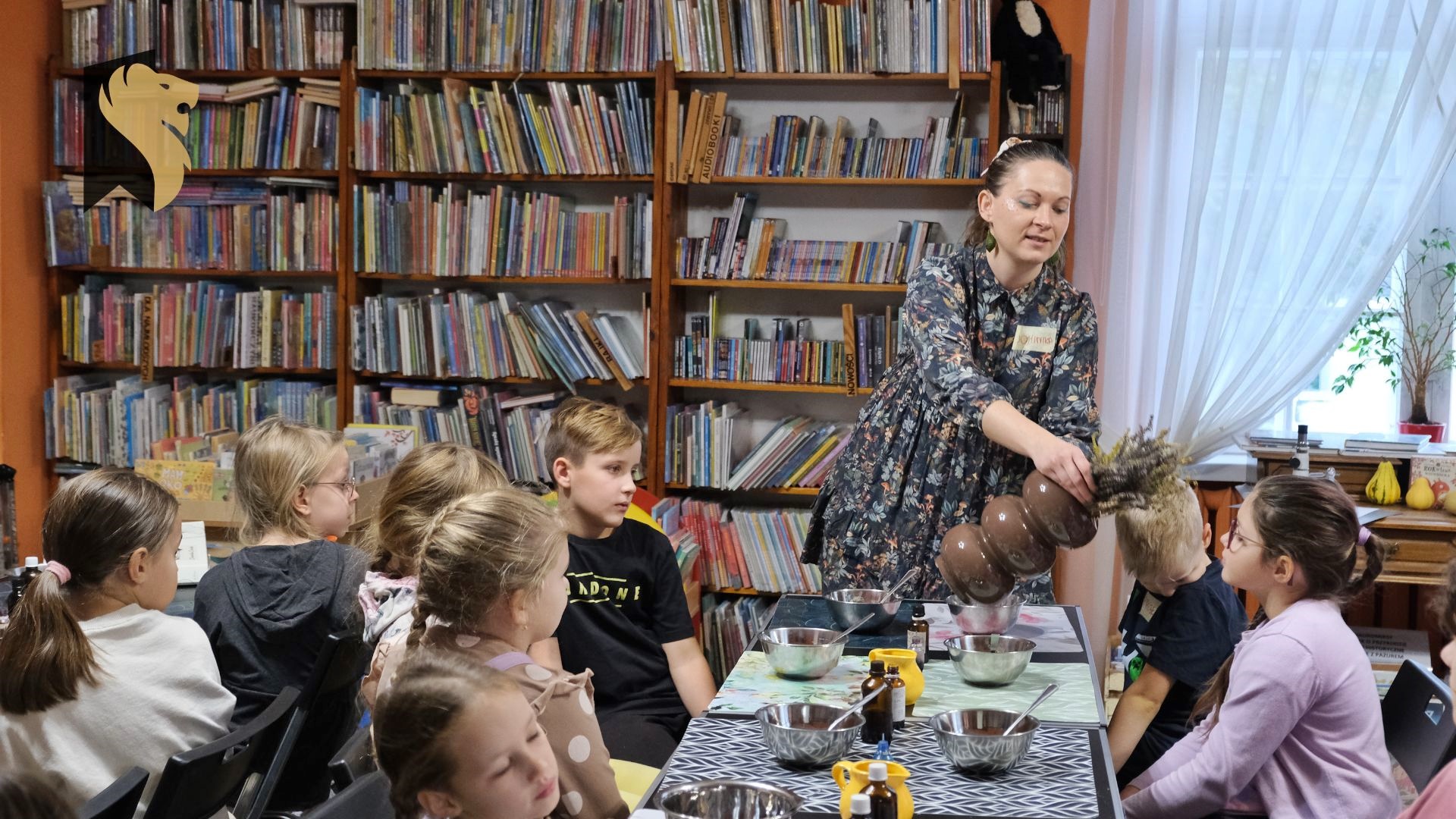 Dzieci siedzą przy stołach w bibliotece, na których leżą kolorowe maty oraz stoją miski. Prowadząca zajęcia- Dominika Dudzic podaje do wąchania zioła.