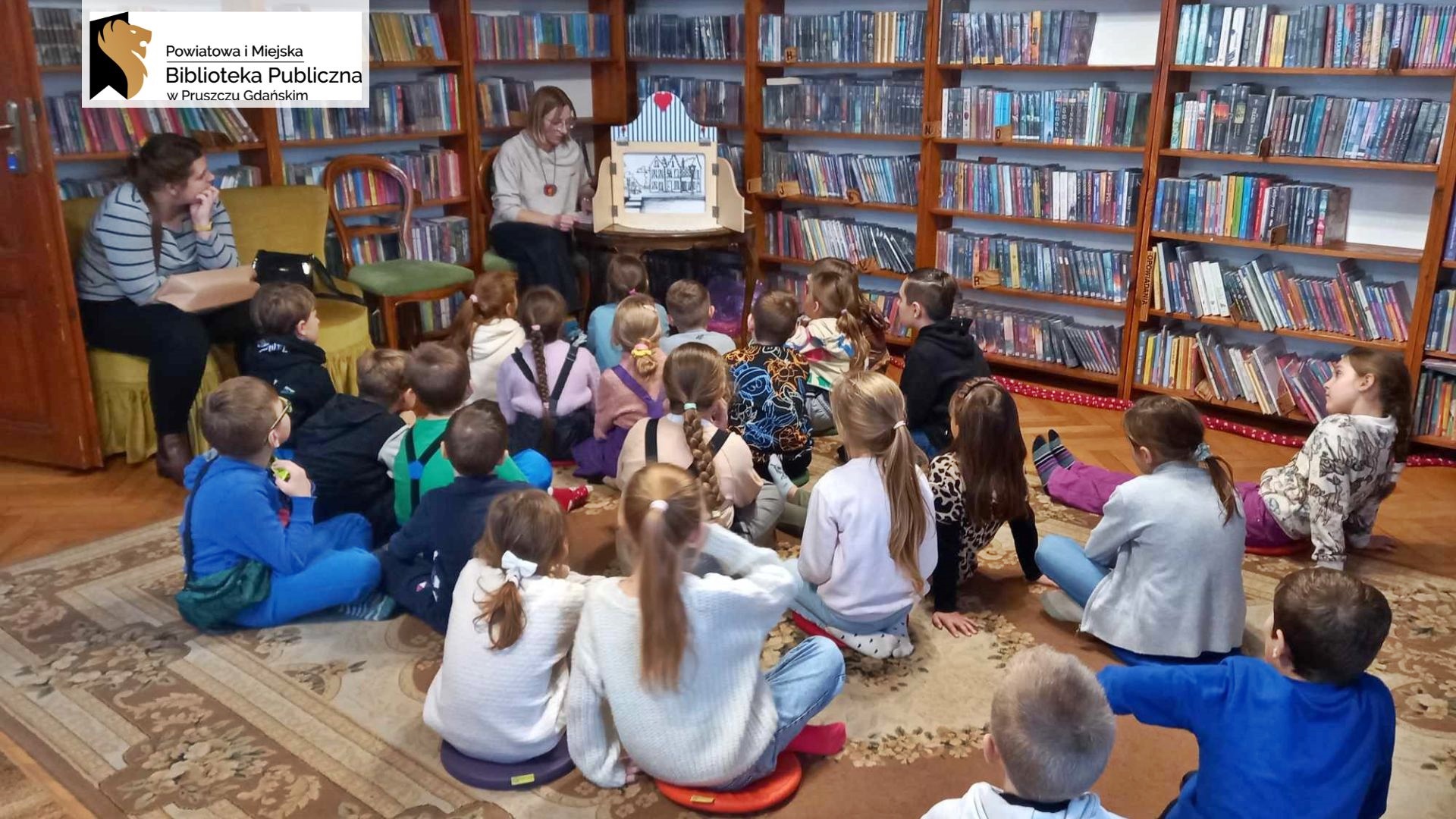 Dzieci siedzą na podłodze i patrzą w kierunku bibliotekarki, która mówi i wykonuje gest dłonią. Po lewej, na stoliku, stoi teatrzyk kamishibai, w środku znajduje się ilustracja. W tle regały z książkami.
