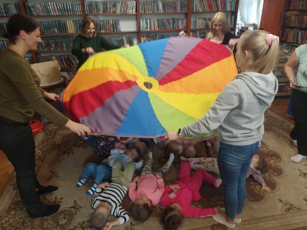 Dzieci leżą na podłodze. Kobiety stoją wokół nich i trzymają chustę klanzy nad głowami dzieci. W tle regały z książkami.