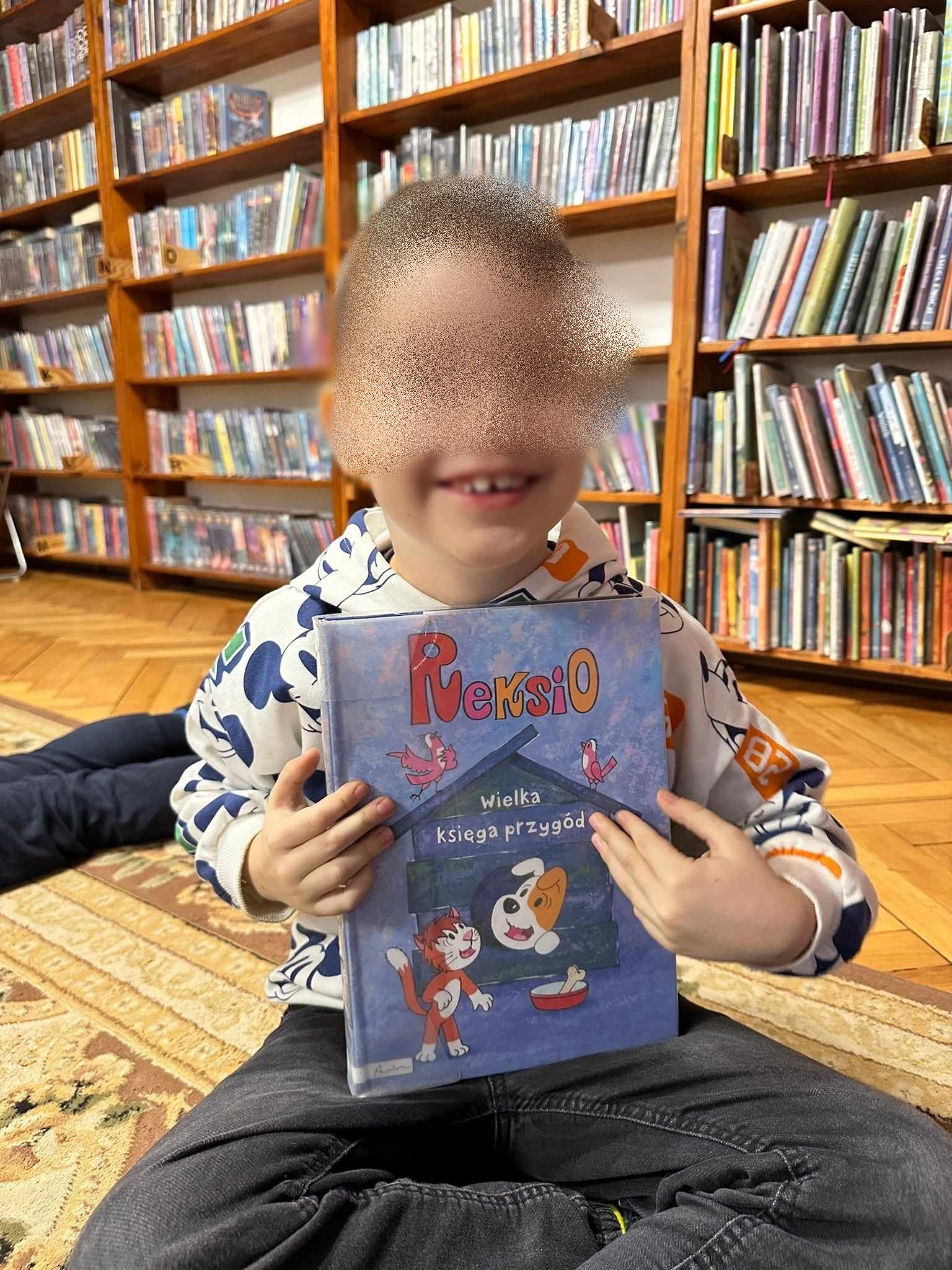 Na dywanie siedzi uśmiechnięty chłopiec, który trzyma w rękach książkę pt. Reksio. Wielka księga przygód.
