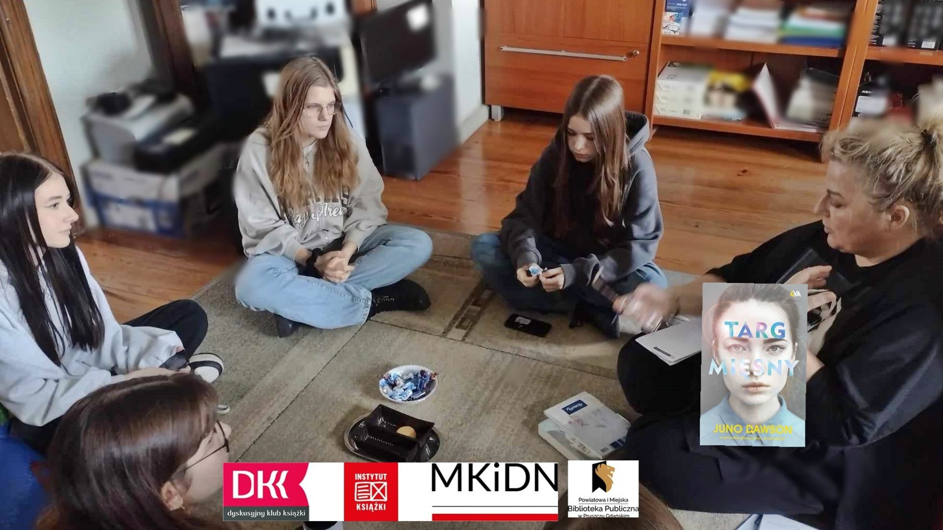 4 dziewczyny z Dyskusyjnego Klubu Książki dla młodzieży siedzą na dywanie. Między nimi siedzi moderatora klubu i coś mówi.