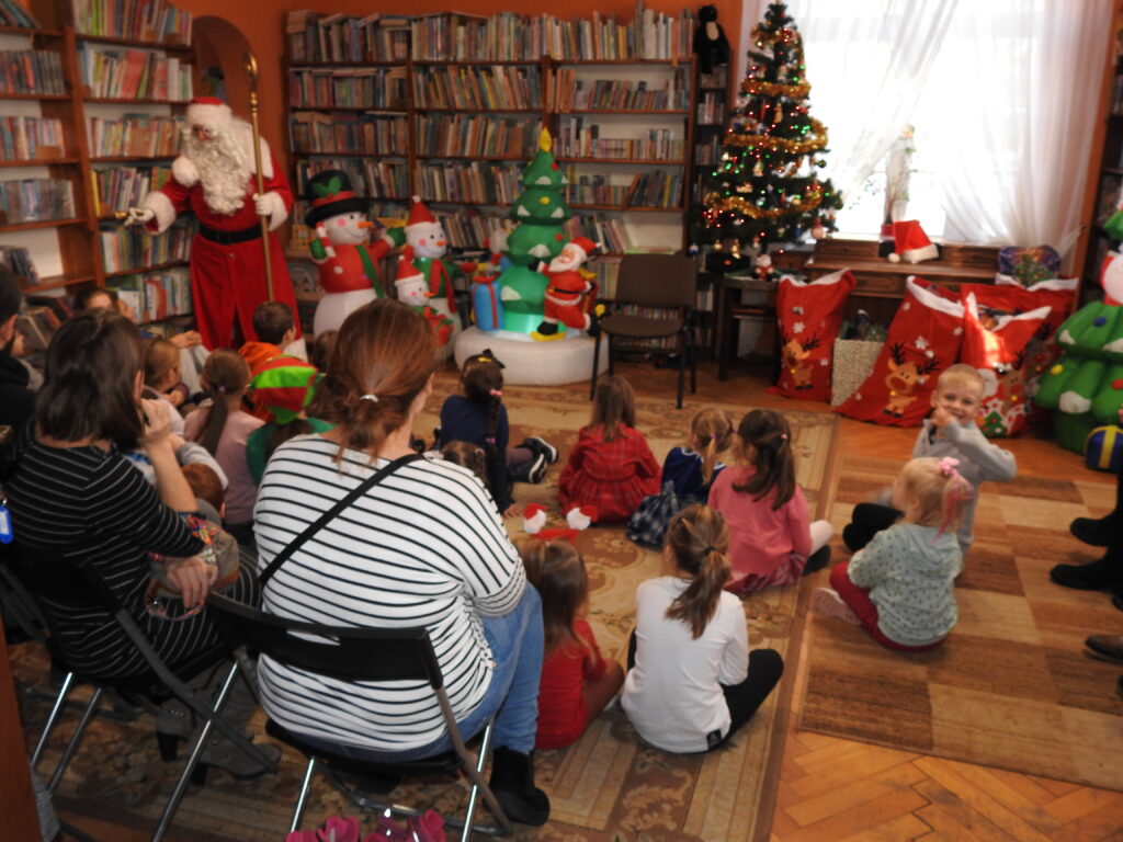  Na dywanie w bibliotece siedzą dzieci i patrzą w stronę mężczyzny w stroju Mikołaja. Po prawej i lewej stronie stoją dmuchane ozdoby świąteczne: choinki, bałwany.
