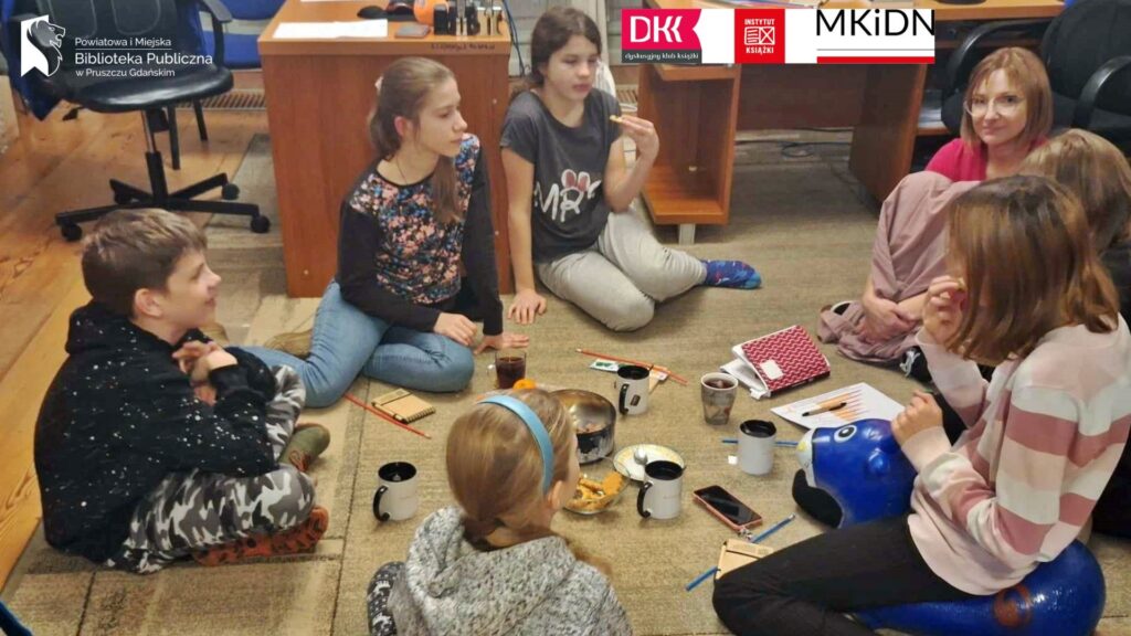 Na dywanie siedzi: 5 dziewczynek, 1 chłopiec oraz moderatorka klubu książki. Pośrodku stoją ciastka w pudełku, kubki z napojem oraz leżą książki.