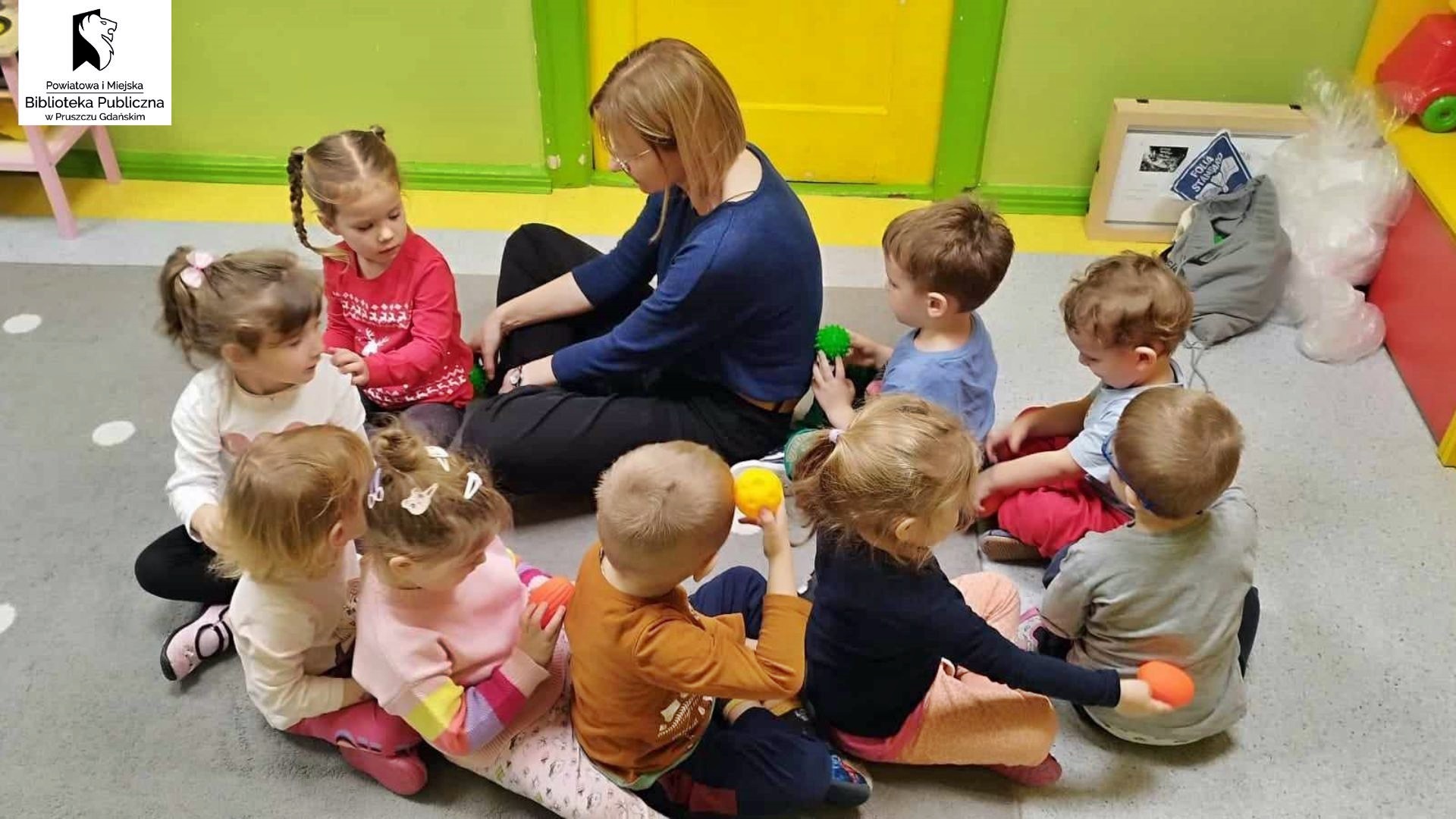 Grupka dzieci oraz bibliotekarka siedzą na dywanie, jedne za drugim. Masują sobie plecy piłeczkami sensorycznymi.